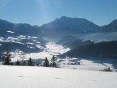 Ein schneebedecktes Tal mit verstreuten Bäumen und Häusern, umgeben von Bergen unter einem klaren blauen Himmel.