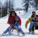 Eine vierköpfige Familie schlittelt einen verschneiten Hügel zwischen blattlosen Bäumen hinunter, alle tragen Winterkleidung und Sonnenbrillen.