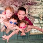 Ein Erwachsener und drei kleine Kinder mit Schwimmbrillen lächeln, während sie in einem Schwimmbad in der Nähe von gefliesten Stufen unter Wasser posieren.