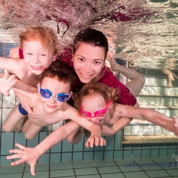 Ein Erwachsener und drei kleine Kinder mit Schwimmbrillen lächeln, während sie in einem Schwimmbad in der Nähe von gefliesten Stufen unter Wasser posieren.