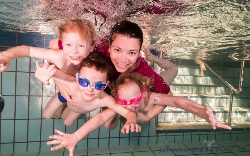 Ein Erwachsener und drei kleine Kinder mit Schwimmbrillen lächeln, während sie in einem Schwimmbad in der Nähe von gefliesten Stufen unter Wasser posieren.