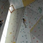 Zwei Personen klettern an einer Indoor-Kletterwand, an der bunte Griffe und Seile zu sehen sind.