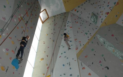 Zwei Personen klettern an einer Indoor-Kletterwand, an der bunte Griffe und Seile zu sehen sind.