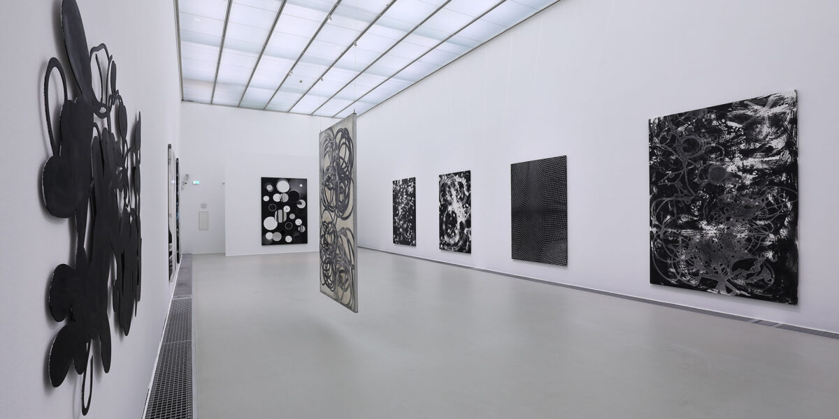 Eine moderne Kunstgalerie mit weißen Wänden zeigt mehrere abstrakte Schwarz-Weiß-Gemälde, darunter zwei hängende Leinwände in der Mitte des Raums.