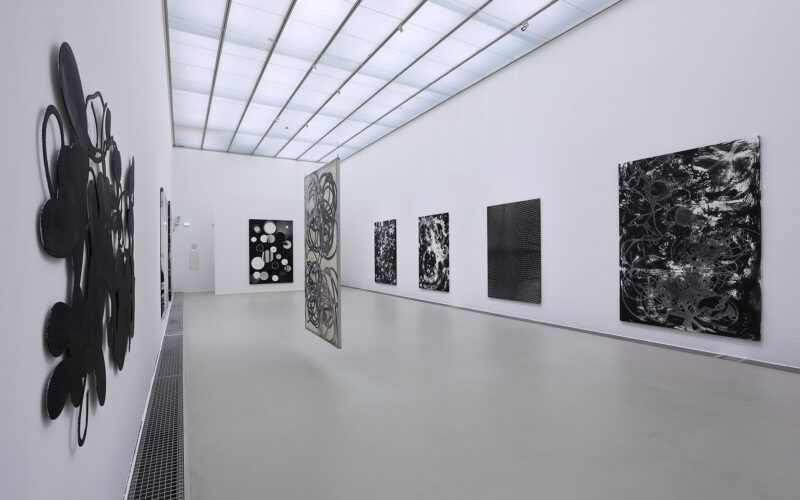 Eine moderne Kunstgalerie mit weißen Wänden zeigt mehrere abstrakte Schwarz-Weiß-Gemälde, darunter zwei hängende Leinwände in der Mitte des Raums.