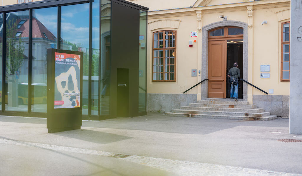 Eine Person betritt ein gelbes Gebäude mit gewölbten Fenstern und einer Glaskonstruktion im Außenbereich. In der Nähe des Eingangs ist ein buntes Plakat angebracht.