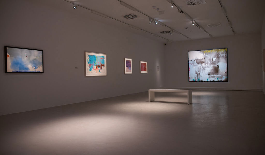 Ein moderner Galerieraum mit abstrakten Gemälden an weißen Wänden, Scheinwerfern darüber und einer weißen Bank in der Mitte.