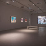 Ein moderner Galerieraum mit abstrakten Gemälden an weißen Wänden, Scheinwerfern darüber und einer weißen Bank in der Mitte.