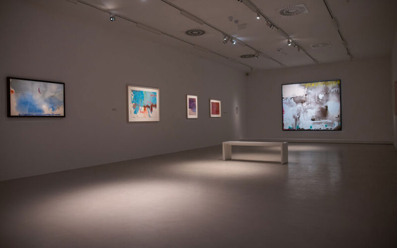 Ein moderner Galerieraum mit abstrakten Gemälden an weißen Wänden, Scheinwerfern darüber und einer weißen Bank in der Mitte.
