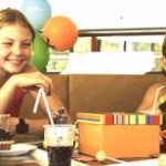 Vier Kinder in Badeanzügen sitzen an einem Tisch, essen und trinken aus Strohhalmen, im Hintergrund sind Luftballons und eine Geschenkbox zu sehen.