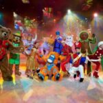 Eine Gruppe von Darstellern in farbenfrohen Tier- und Figurenkostümen posiert gemeinsam auf einer hell erleuchteten Bühne mit einem festlichen Hintergrund.