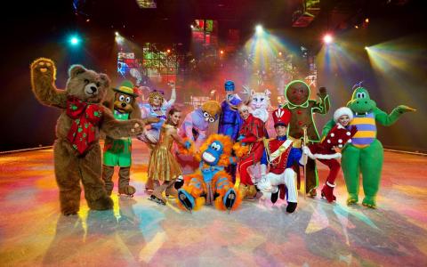 Eine Gruppe von Darstellern in farbenfrohen Tier- und Figurenkostümen posiert gemeinsam auf einer hell erleuchteten Bühne mit einem festlichen Hintergrund.