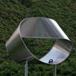 Eine große metallene Möbiusband-Skulptur steht im Freien vor einem grünen, begrünten Hintergrund.