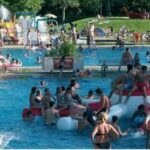 Ein überfülltes öffentliches Freibad mit Menschen, die bei sonnigem Wetter schwimmen, faulenzen und auf einer großen aufblasbaren Struktur spielen.