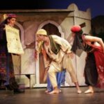 Vier Schauspieler in farbenfrohen, historischen Kostümen spielen auf der Bühne eine dramatische Szene, wobei einer eine Schriftrolle hält und die anderen vor einer gemalten Gebäudekulisse lehnen.