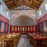 Großer, hoher Raum mit Holztischen und -stühlen, rot und schwarz gestrichenen Wänden, gewölbter Steinnische und auf Tischen angeordneten Speisen.