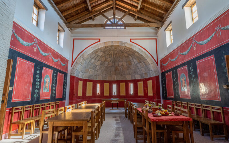 Großer, hoher Raum mit Holztischen und -stühlen, rot und schwarz gestrichenen Wänden, gewölbter Steinnische und auf Tischen angeordneten Speisen.