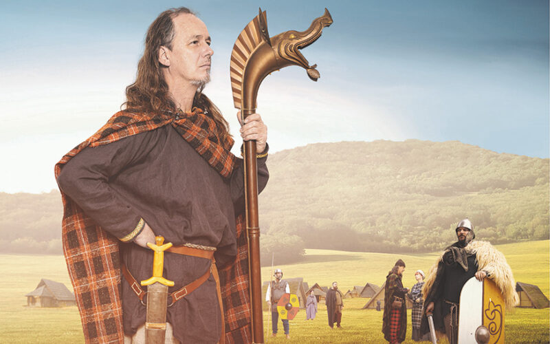 Ein Mann in historischer Kleidung und mit einem Stab in der Hand steht auf einem Feld mit anderen Personen in ähnlicher Kleidung und mit Schilden, mit Zelten und Hügeln im Hintergrund.