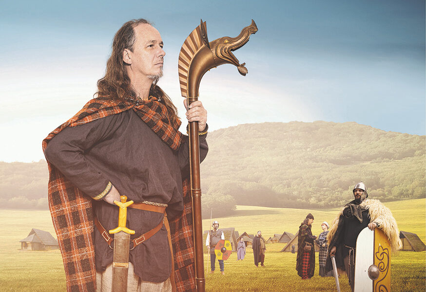 Ein Mann in historischer Kleidung und mit einem Stab in der Hand steht auf einem Feld mit anderen Personen in ähnlicher Kleidung und mit Schilden, mit Zelten und Hügeln im Hintergrund.