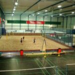 Sporthalle mit einem Sandvolleyballfeld, das von einem Zaun umgeben ist, und weiteren Feldern und Geräten im Vordergrund.