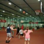 Eine Gruppe von Kindern spielt drinnen auf einem Platz Basketball. Ein Kind wirft den Ball in Richtung des Korbes, während die anderen zuschauen oder versuchen, ihn zu blocken.