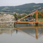 Eine Holzbrücke überquert ein ruhiges Gewässer mit einer felsigen Struktur und einer großen rot-weiß senkrecht gestreiften Flagge in der Nähe; im Hintergrund sind Hügel und Häuser zu sehen.