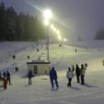 Menschen fahren Ski und stehen in der Nähe eines Skilifts auf einer verschneiten, beleuchteten Skipiste am Abend mit Bäumen im Hintergrund.