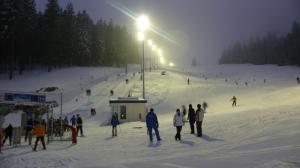 Menschen fahren Ski und stehen in der Nähe eines Skilifts auf einer verschneiten, beleuchteten Skipiste am Abend mit Bäumen im Hintergrund.