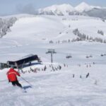 Ein Skifahrer in einer roten Jacke fährt einen verschneiten Hang in der Nähe eines Skilifts hinunter, mit anderen Skifahrern und schneebedeckten Bergen im Hintergrund.