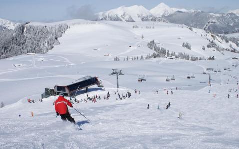 Ein Skifahrer in einer roten Jacke fährt einen verschneiten Hang in der Nähe eines Skilifts hinunter, mit anderen Skifahrern und schneebedeckten Bergen im Hintergrund.