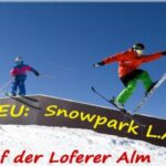 Zwei Skifahrer in farbenfroher Ausrüstung springen von einer Rampe im Snowpark L.A., Loferer Alm, unter blauem Himmel. Ein Text in deutscher Sprache wirbt für den neuen Standort des Snowparks.