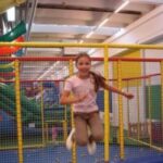Ein junges Mädchen springt auf einem Trampolin in einem Indoor-Spielbereich mit bunten Kletterstrukturen und Sicherheitsnetzen.