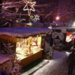 Die verschneiten Marktstände unter freiem Himmel sind nachts mit Lichterketten und Sternen beleuchtet, und zwischen den Ständen laufen ein paar Leute auf einem verschneiten Weg.