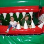 Zwei Kinder sitzen und spielen auf einer aufblasbaren Struktur, die wie das offene Maul eines großen grünen Wesens mit weißen spitzen Zähnen aussieht.