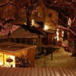 Ein verschneiter Markt im Freien bei Nacht, beleuchtet von Lichterketten und dekorierten Ständen, mit kahlen Bäumen und schneebedecktem Boden.