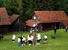 Kinder spielen und klettern auf Holzkonstruktionen auf einer Wiese in der Nähe von rustikalen Holzhäusern mit roten Ziegeldächern, umgeben von Bäumen.