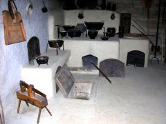 Historische Küche mit weiß getünchten Wänden, alten Kochtöpfen und Öfen; ein Holzschemel und eine offene Kellerluke auf dem Steinboden sind zu sehen.