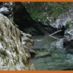 Ein kleiner Bach fließt zwischen großen Felsen und Geröll, gesäumt von Grün und zerklüftetem Terrain.