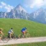 Vier Personen fahren mit ihren Fahrrädern auf einem Schotterweg durch ein grasbewachsenes Feld mit hohen, felsigen Bergen im Hintergrund unter einem teilweise bewölkten Himmel.