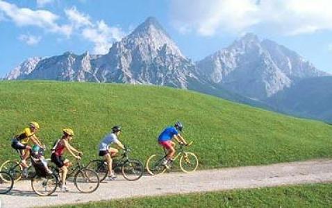 Vier Personen fahren mit ihren Fahrrädern auf einem Schotterweg durch ein grasbewachsenes Feld mit hohen, felsigen Bergen im Hintergrund unter einem teilweise bewölkten Himmel.