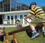 Zwei Kinder spielen auf einer hölzernen Wippe im Freien in der Nähe eines modernen Gebäudes, mit Bergen im Hintergrund.