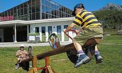 Zwei Kinder spielen auf einer hölzernen Wippe im Freien in der Nähe eines modernen Gebäudes, mit Bergen im Hintergrund.
