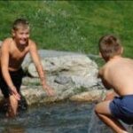 Drei Jungen in Badehosen spielen und planschen in einem seichten Bach im Freien, mit Gras und Felsen im Hintergrund.