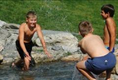 Drei Jungen in Badehosen spielen und planschen in einem seichten Bach im Freien, mit Gras und Felsen im Hintergrund.