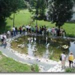 Eine Gruppe von Menschen steht am Rande eines kleinen Teichs, umgeben von Gras, Bäumen und einigen Gebäuden im Hintergrund.