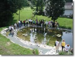 Eine Gruppe von Menschen steht am Rande eines kleinen Teichs, umgeben von Gras, Bäumen und einigen Gebäuden im Hintergrund.