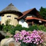 Ein rundes, strohgedecktes Gebäude mit schrägem Dachausbau, umgeben von Grün und blühenden rosa Blumen im Vordergrund.