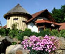 Ein rundes, strohgedecktes Gebäude mit schrägem Dachausbau, umgeben von Grün und blühenden rosa Blumen im Vordergrund.