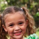 Ein junges Mädchen mit lockigem Haar in Zöpfen lächelt, während es einen grün-weiß gestreiften Gegenstand im Freien hält.