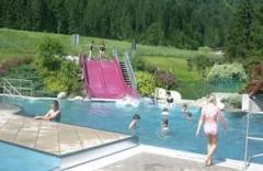 Kinder spielen und schwimmen in einem Außenpool mit einer rosafarbenen Doppelrutsche, umgeben von Grün und Bäumen im Hintergrund.